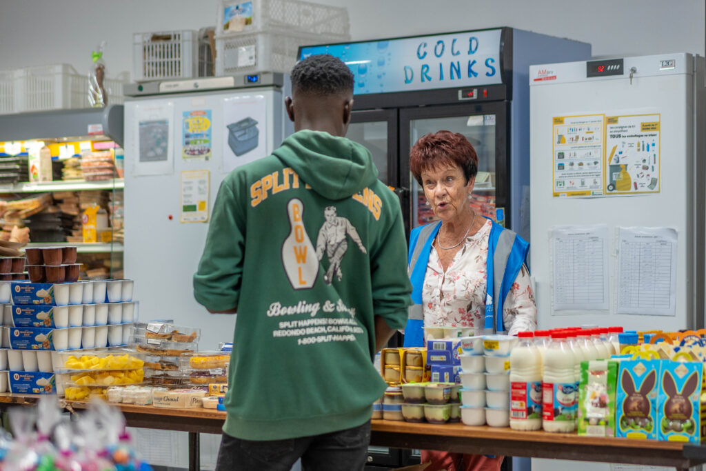 Une bénévole discute avec un jeune homme bénéficiaire de l'aide alimentaire autour des denrées proposées.